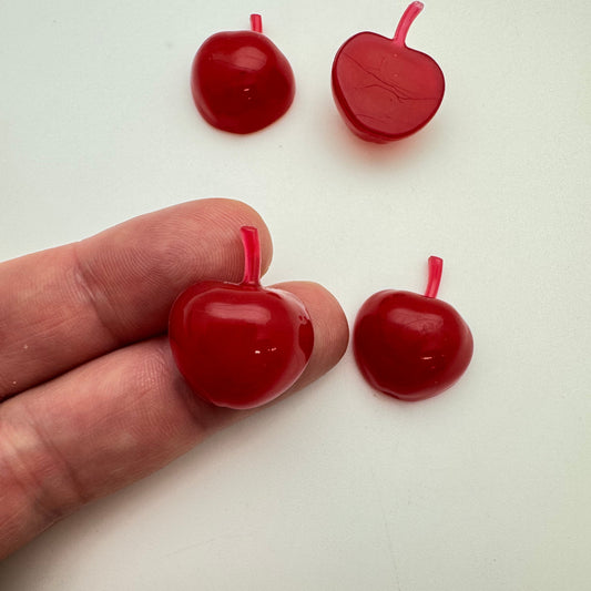 Red resin cabachons in the shape of an apple on a white background with a hand holding one of them.