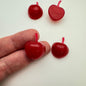 Red resin cabachons in the shape of an apple on a white background with a hand holding one of them.