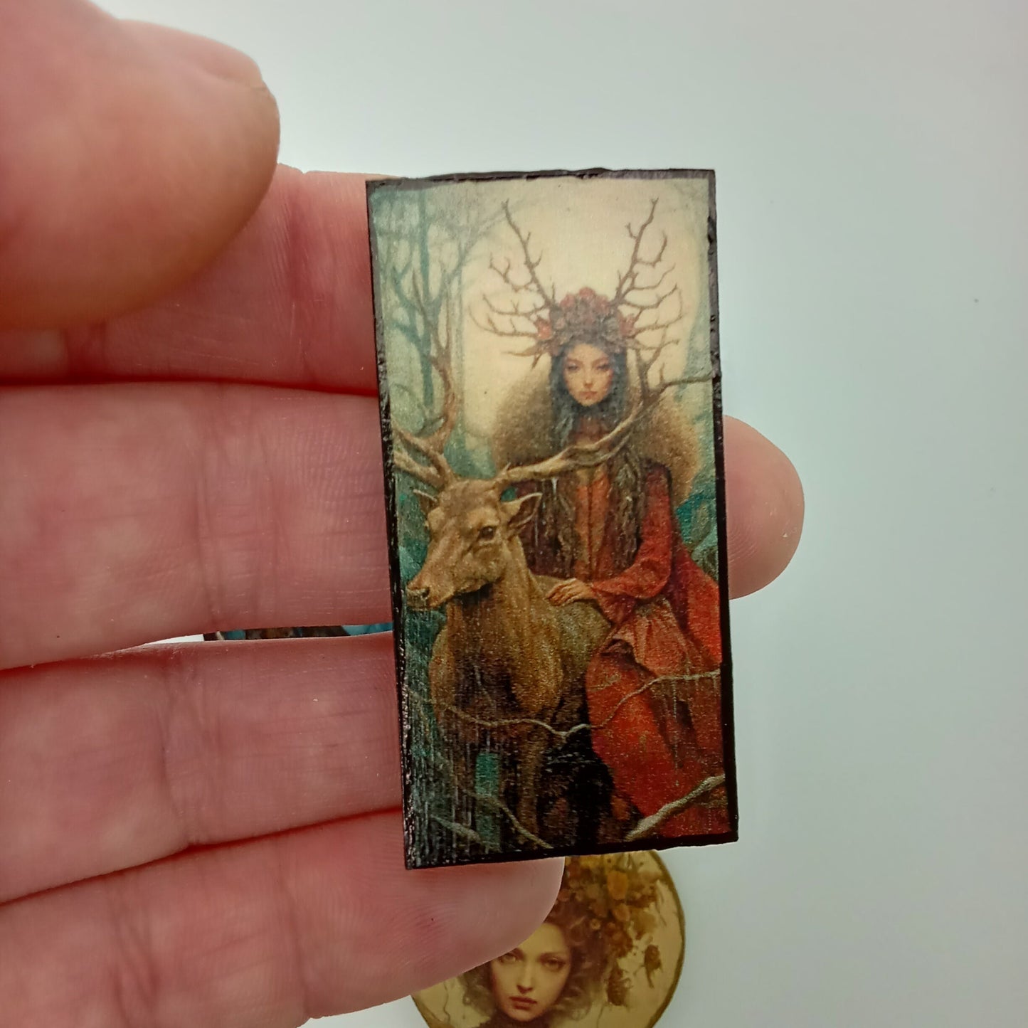 Small rectangular artwork held between fingers with a coin for scale on a white background