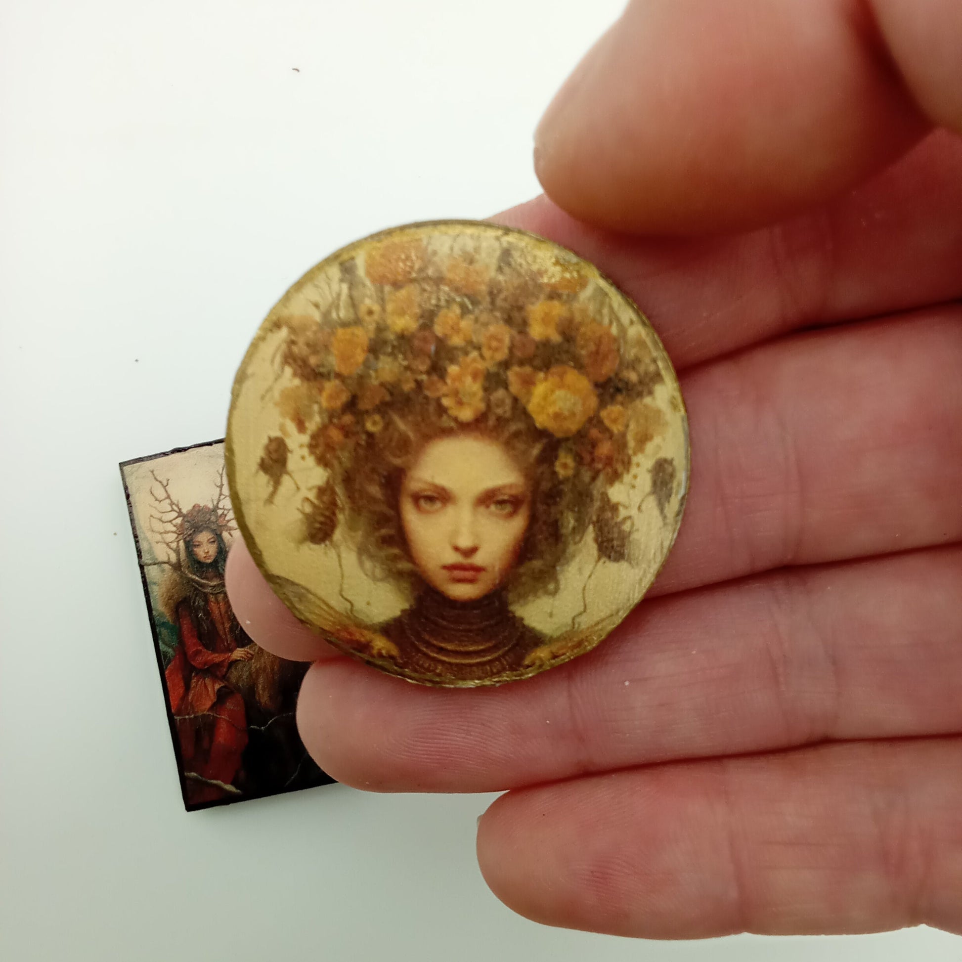 Round decorative wood cabachon with a face design held by a hand on a white background